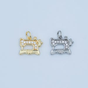 May include: Two sewing machine charms. One is gold-colored, the other is silver-colored. Both charms have a row of clear, round stones across the top. The charms are detailed with small knobs and a decorative base. The background is a light, solid color.