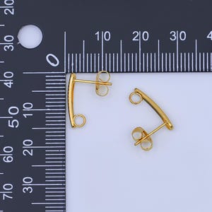 May include: Two gold-colored, curved bar earrings with a post and loop design. The earrings are shown against a white background with a black ruler for scale. The earrings are small and delicate.