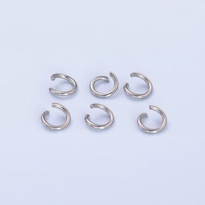 May include: Six silver jump rings, also known as split rings, are arranged in a staggered pattern on a light blue background.