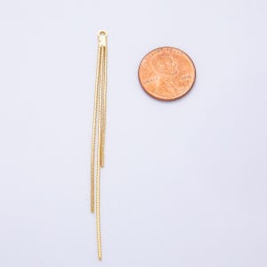 May include: Gold-colored jewelry component with three dangling chains. The component has a small loop at the top for attachment. A penny is next to the jewelry for size comparison. The chains are thin and have a textured appearance.
