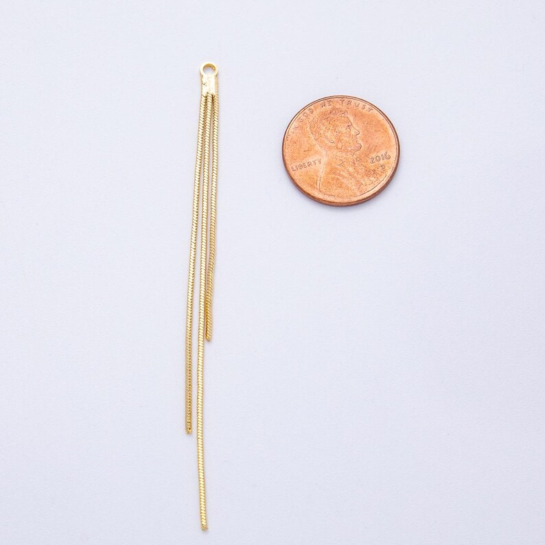 May include: Gold-colored jewelry component with three dangling chains. The component has a small loop at the top for attachment. A penny is next to the jewelry for size comparison. The chains are thin and have a textured appearance.