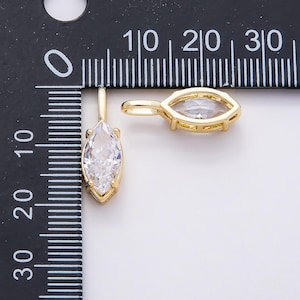 Op de afbeelding: Twee goudkleurige hangerbedels met een marquise-geslepen, heldere kristal. De hangers worden getoond naast een liniaal, waarbij het kristal ongeveer 1,3 cm lang is. De bedels hebben een eenvoudig, elegant ontwerp.