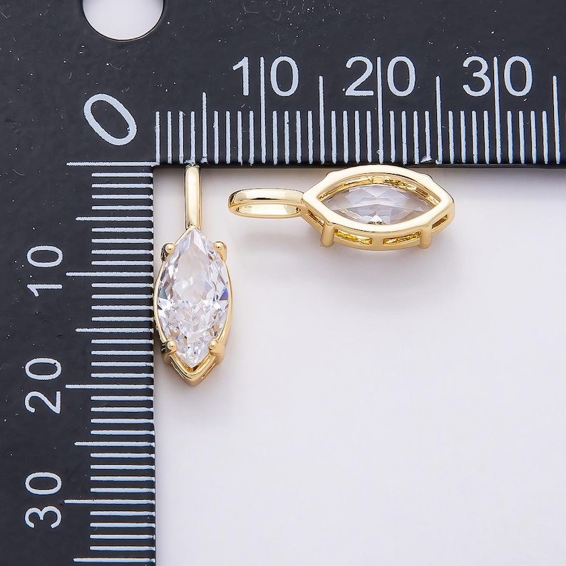 Op de afbeelding: Twee goudkleurige hangerbedels met een marquise-geslepen, heldere kristal. De hangers worden getoond naast een liniaal, waarbij het kristal ongeveer 1,3 cm lang is. De bedels hebben een eenvoudig, elegant ontwerp.