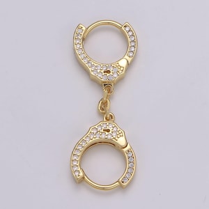 May include: Gold-colored hoop earrings in a handcuff design. Each earring features a circular hoop with a decorative handcuff-shaped charm. The charms are embellished with small, clear crystals, adding sparkle to the jewelry.