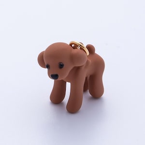 Puede incluir: Una figura de perro marrón con un anillo dorado sujeto a su espalda. El perro tiene un acabado liso y mate, con pequeños ojos negros y una nariz negra. El perro está de pie sobre sus cuatro patas.