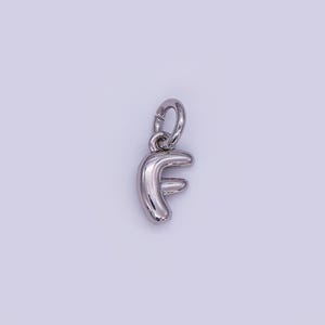 May include: A silver-colored charm in the shape of the letter "F". The charm has a rounded, three-dimensional design and includes a small loop at the top for attachment to a necklace or bracelet. The background is a light purple color.