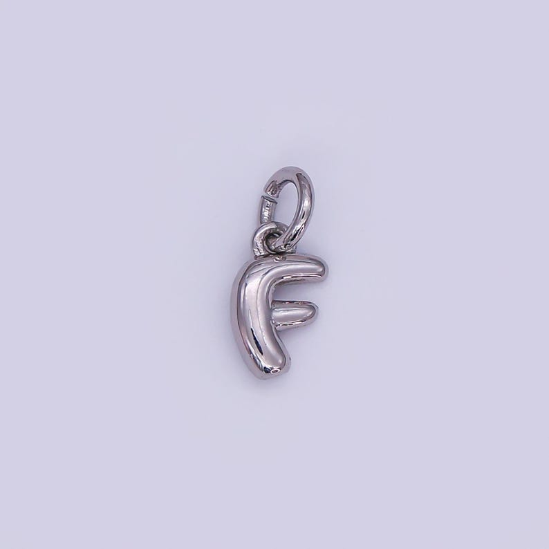 May include: A silver-colored charm in the shape of the letter "F". The charm has a rounded, three-dimensional design and includes a small loop at the top for attachment to a necklace or bracelet. The background is a light purple color.