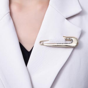 May include: A gold-colored safety pin brooch with a row of small, dark stones set along the curved end. The brooch is pinned to a white blazer, creating a stylish accessory. The pin is a unique piece of jewelry.