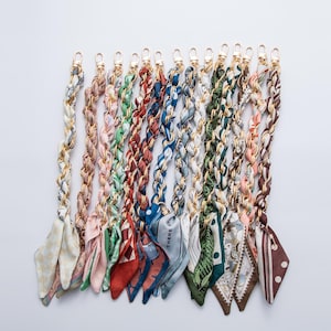 May include: A collection of colorful purse straps with gold-tone chain links and patterned silk scarf accents. The straps feature various colors and patterns, including floral, geometric, and abstract designs. Each strap has a clasp for easy attachment.