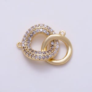 May include: A gold-toned jewelry connector featuring two interlocking circular rings. One ring is encrusted with numerous small, clear, sparkling cubic zirconia stones, while the other ring is a smooth, polished gold-toned metal band.