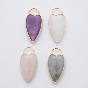 May include: Four gemstone pendants in heart shapes with gold-colored metal frames and loops. The gemstones are amethyst, white quartz, rose quartz, and gray labradorite. The pendants are displayed against a white background.