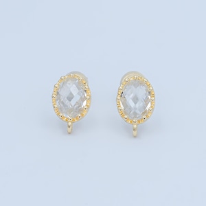 May include: A pair of gold-colored stud earrings. Each earring features a clear, faceted, oval-shaped stone set within a beaded gold frame. The earrings have a small loop at the bottom for attaching to a chain or other jewelry component.