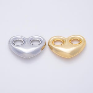 May include: Two heart-shaped charms, one in a polished silver tone and one in a polished gold tone. Each heart features two circular openings at the top, creating a smooth, rounded, and metallic surface with a reflective finish on both pieces.