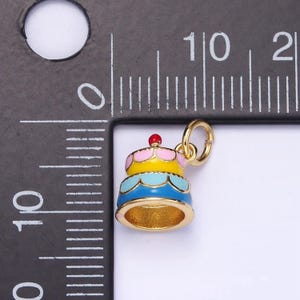 May include: A gold-colored charm in the shape of a three-tiered cake with pink, yellow, blue, and light blue frosting. The charm has a red cherry on top and a gold jump ring.