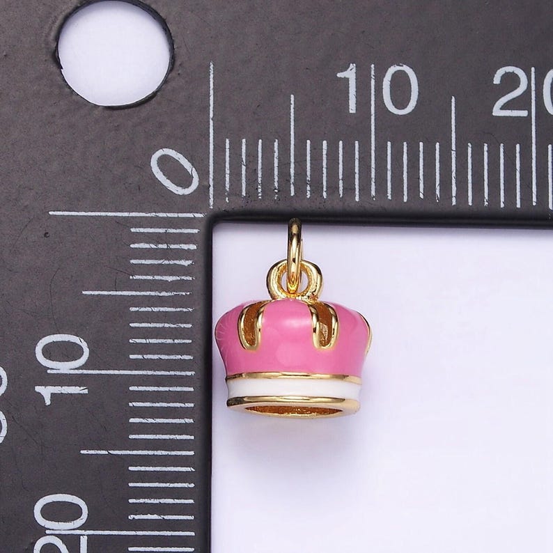 May include: A pink and gold crown charm with a white band. The charm is about 1 cm in diameter.