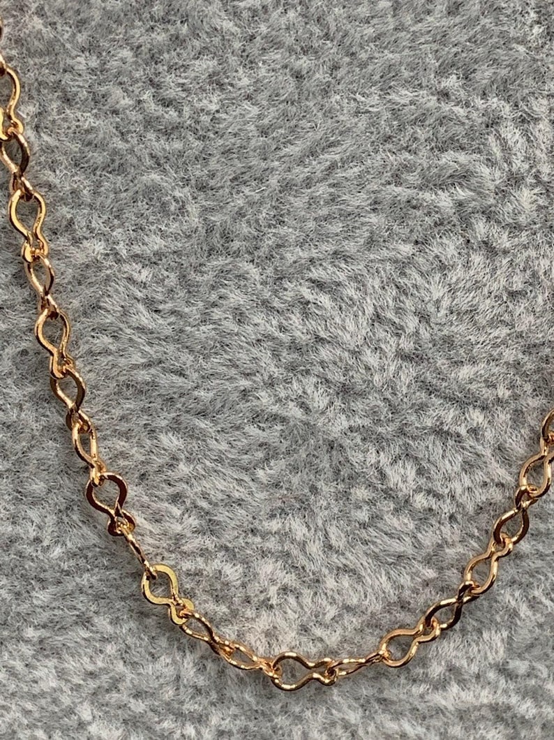 Op de afbeelding: Een gouden ketting met een delicaat, gekoppeld ontwerp. De ketting is gemaakt van kleine, met elkaar verbonden lussen die een vloeiende, elegante look cre&euml;ren.