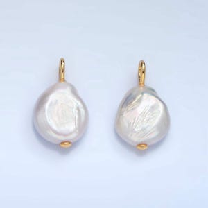 Può includere: Due orecchini con perle barocche bianche con elementi dorati. Le perle hanno una forma irregolare e organica e una superficie lucida. Gli orecchini sono esposti su uno sfondo chiaro.