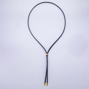 K&ouml;nnte beinhalten: Eine schwarze Kordelkette mit einem goldfarbenen Metallakzent und zwei goldfarbenen Metallspitzen. Die Halskette wird vor einem wei&szlig;en Hintergrund pr&auml;sentiert.