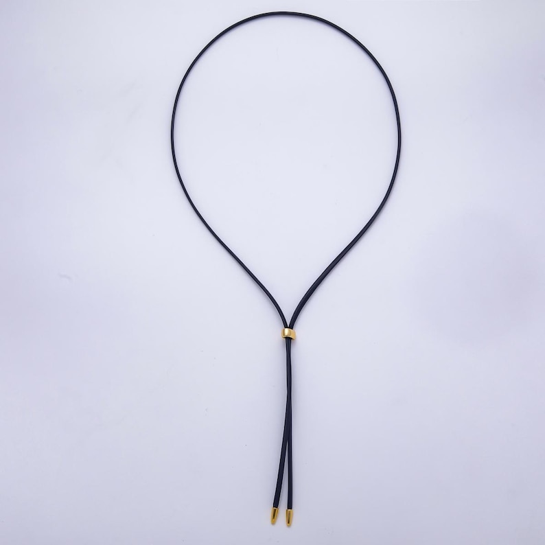 K&ouml;nnte beinhalten: Eine schwarze Kordelkette mit einem goldfarbenen Metallakzent und zwei goldfarbenen Metallspitzen. Die Halskette wird vor einem wei&szlig;en Hintergrund pr&auml;sentiert.