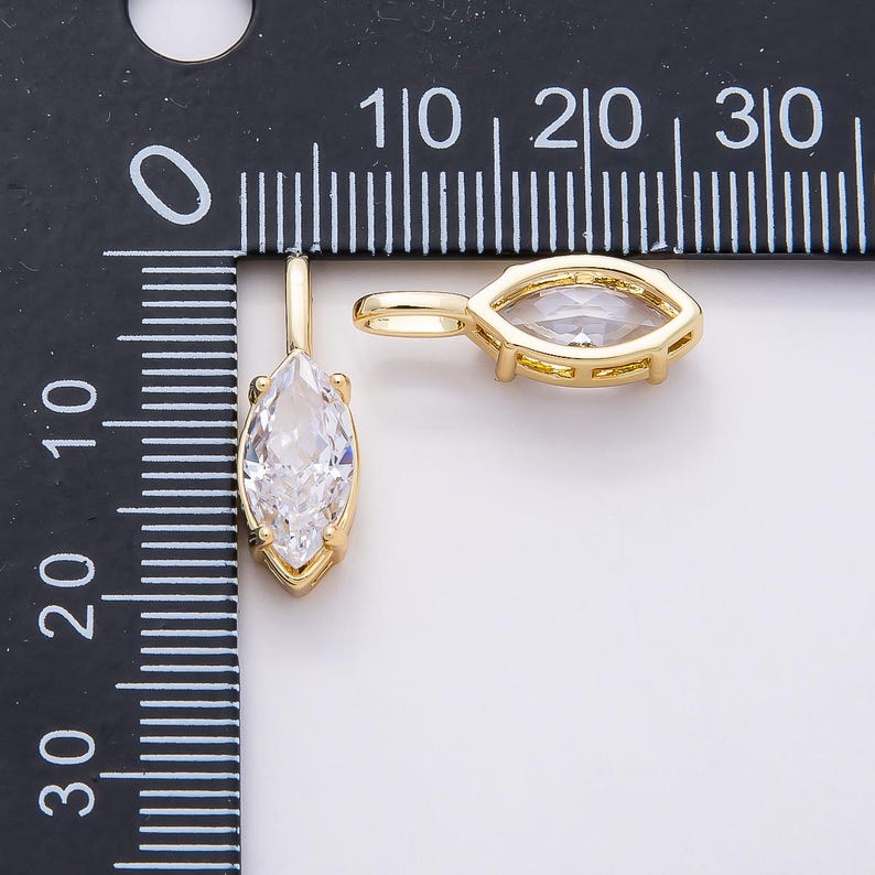 Op de afbeelding: Twee goudkleurige hangerbedels met een marquise-geslepen, helder kristal. De hangers worden weergegeven tegen een witte achtergrond met een zwarte liniaal voor de schaal. Het kristal is ongeveer 1,9 cm lang.