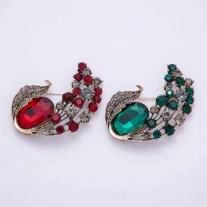 May include: Two decorative brooches with a leaf design. One brooch features a large red gemstone and red, gray, and clear accent stones. The other brooch has a large green gemstone and green, gray, and clear accent stones. Both have gold-colored metal and pin backs.