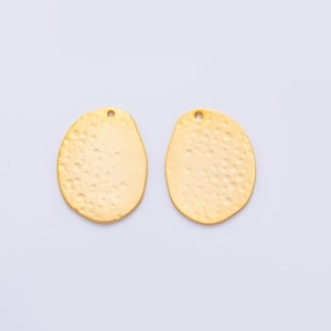 May include: Two gold-colored, hammered-texture oval pendants with a small hole at the top for stringing. The pendants have an irregular shape and a slightly rough surface, suggesting a handmade or artisan quality. The background is plain white.