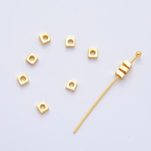 May include: Gold-colored square beads with a hole in the center, scattered on a white surface. A gold-colored beading needle with three square beads is also present. These beads are ideal for jewelry making.