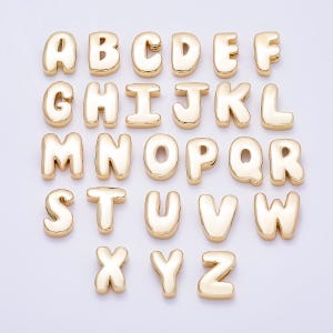 May include: A collection of twenty-six individual gold-colored metal alphabet letter charms, arranged in rows from A to Z. Each letter features a smooth, rounded, and puffed surface with a reflective finish, designed as individual pieces.