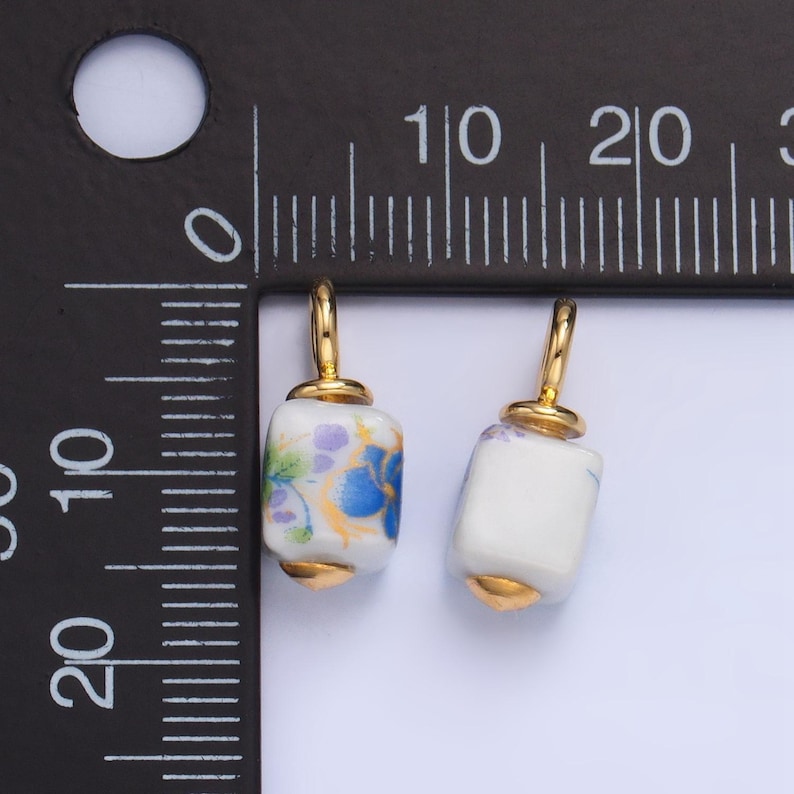 May include: Two white ceramic square beads with gold accents and floral patterns. The beads have a gold loop at the top for hanging.
