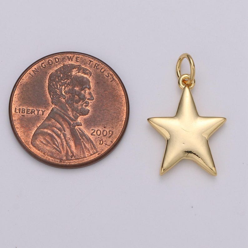 Puede incluir: Un dije de estrella en tono dorado con una peque&ntilde;a argolla unida a la punta superior. El dije est&aacute; colocado junto a una moneda de cobre de un centavo de los Estados Unidos para comparar el tama&ntilde;o, con el perfil de Abraham Lincoln y el a&ntilde;o 2009.