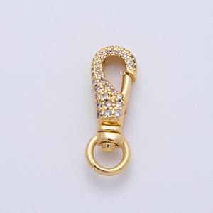 May include: A gold-coloured lobster clasp with a ring at the bottom and a curved top encrusted with small, clear gemstones. The clasp is designed for jewellery making or attaching to accessories.