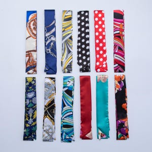 Könnte beinhalten: Eine Sammlung von zwölf farbenfrohen Seidenschals, jeder mit einem einzigartigen Muster. Designs umfassen Polka-Punkte, Blumendrucke und geometrische Formen in verschiedenen Farben wie Rot, Blau, Schwarz und Gelb. Die Schals sind rechteckig und aus glänzendem Stoff.