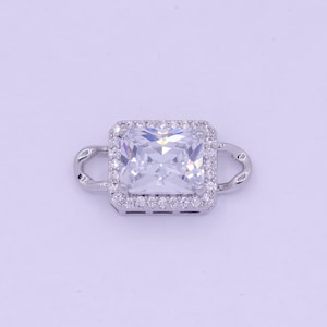 May include: A silver charm with a large, clear square-shaped cubic zirconia stone surrounded by smaller stones. The charm has two loops on the sides.