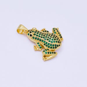 May include: A gold-colored frog pendant with emerald green gemstones covering the body. The frog has black eyes and is set against a white background. The pendant has a loop for a chain.