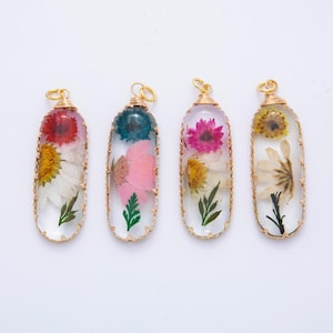Puede incluir: Cuatro colgantes ovalados de resina transparente, cada uno con una flor seca en un marco dorado. Las flores son de color rojo, rosa, amarillo y blanco. Cada colgante tiene un lazo de alambre dorado para colgar.