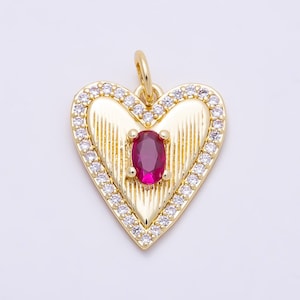 May include: A gold-toned heart-shaped pendant featuring a central oval-cut deep pink gemstone. The surface of the heart has vertical ribbed detailing and is bordered by a frame of small, clear, round-cut white stones. A small gold jump ring is attached.