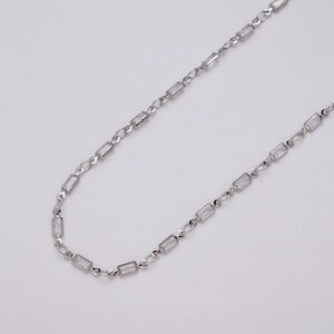 May include: A silver-toned necklace featuring rectangular, clear crystal accents. The chain is delicate, with small links connecting the crystal elements. The necklace is displayed against a plain, light background, highlighting its simple elegance.