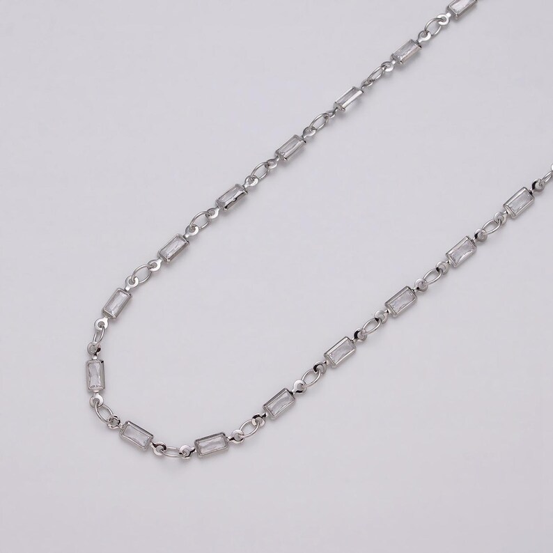 May include: A silver-toned necklace featuring rectangular, clear crystal accents. The chain is delicate, with small links connecting the crystal elements. The necklace is displayed against a plain, light background, highlighting its simple elegance.