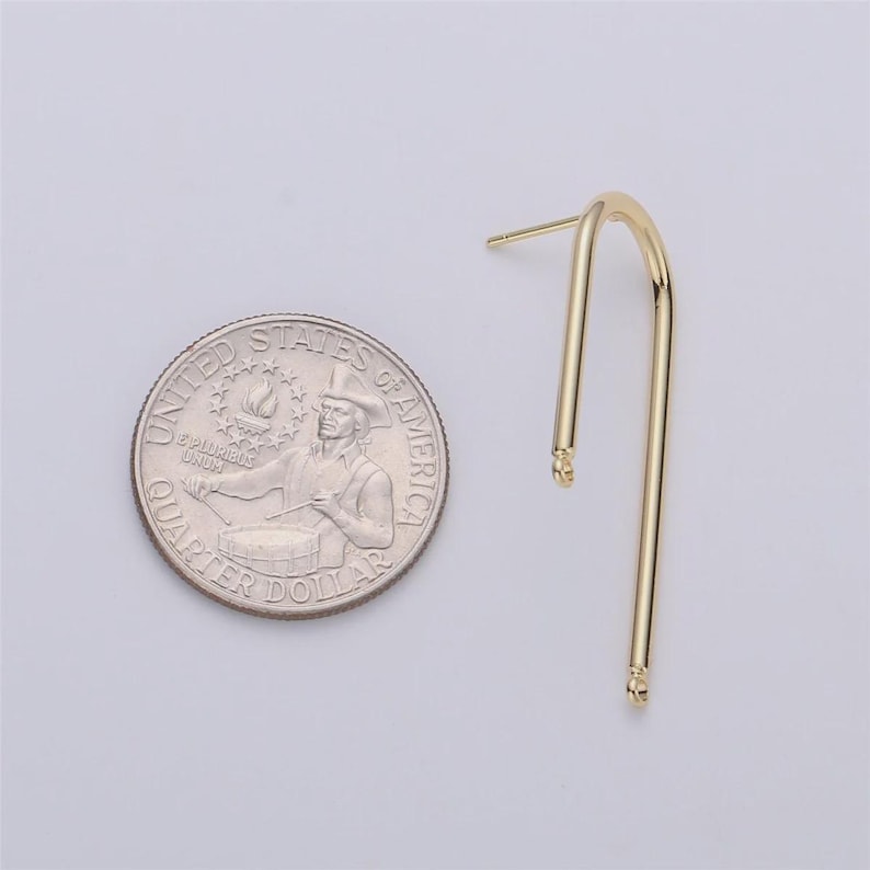 May include: A single gold-colored earring with a curved, elongated design. The earring is next to a U.S. quarter coin for size comparison. The earring has a post for insertion and a small loop at the bottom.