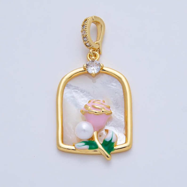 May include: Gold-tone pendant with a rectangular frame, featuring a pink rose, a white pearl, and green leaves. The pendant has a clear gem and a loop adorned with small clear gems. The background is a pearlescent white.