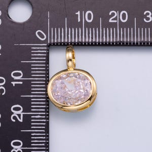 May include: A gold-toned oval pendant featuring a faceted light pink stone set in a polished metal frame. The pendant is positioned against a black measuring tool, showing a width of approximately 0.5 inches and a height of 0.5 inches.