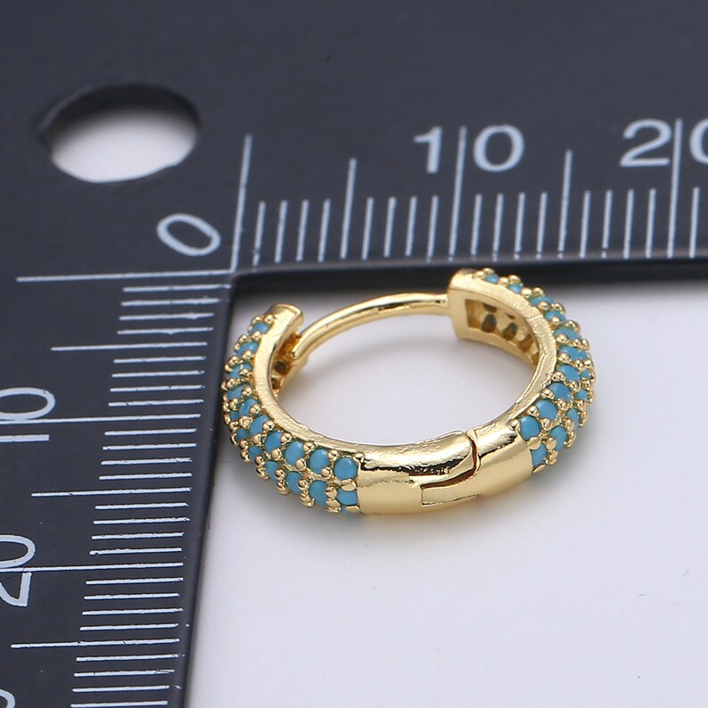 Pu&ograve; includere: Un orecchino a cerchio color oro con una fila di piccole pietre color turchese. L'orecchino &egrave; mostrato accanto a un righello, con un diametro di circa 1,3 cm.