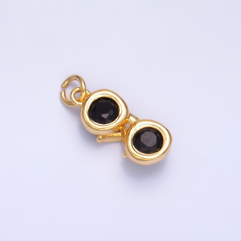 May include: A gold-colored charm in the shape of sunglasses. The charm features two round black stones for lenses. The frame and the loop for attaching the charm are gold-toned. The charm is small and detailed.