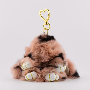 May include: A plush bunny keychain in shades of pink and black, featuring white leather-like paws with gold stitching. The keychain has a gold heart-shaped clasp.