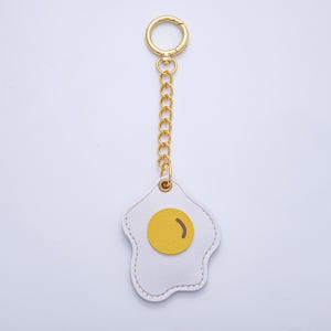May include: A white leather keychain in the shape of a fried egg with a yellow yolk and a gold-colored chain and clasp. The keychain is a fun and quirky accessory.