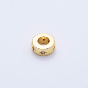 May include: A gold-colored, cylindrical bead with a central hole. The bead is adorned with star-shaped designs, each featuring a small, pink gemstone at its center. The surface has a polished finish, reflecting light.