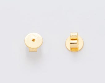 May include: Two gold-colored earring backs on a white background. One back is a flat disc with a post, and the other is a butterfly clutch style. The earring backs are made of a shiny, metallic material.