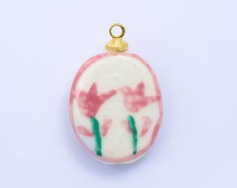 May include: Oval-shaped pendant with a gold-colored loop. The pendant features a white background with a pink border and two pink flower illustrations with green stems. The pendant is likely ceramic or porcelain.