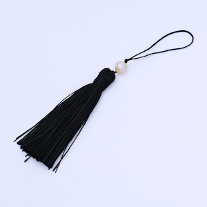 May include: A black tassel with a loop and a white pearl bead. The tassel is made of many strands of black thread, and the pearl is round and smooth. The loop is made of black thread.