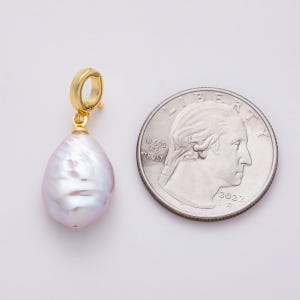 May include: A gold-colored earring pendant with a large, pear-shaped, iridescent, light purple pearl. The pendant is next to a U.S. quarter coin for size comparison. The coin shows the year 2022.
